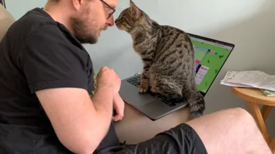 Side on view of a white man with short hair and a beard wearing glasses, black t-shirt, and black shorts. He’s nose to nose with a brown mottled kitten, who is sitting on a laptop keyboard almost completely obscuring the screen.