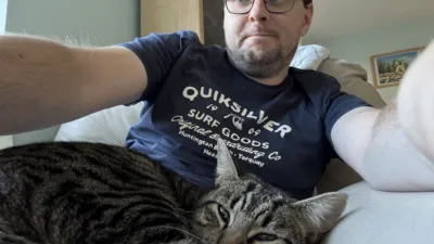 A mottled brown cat looks into the camera from the foreground. In the background is a middle aged man looking unimpressed by the cat’s decision to curl up more when asked to move.