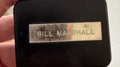 A black tape measure with a brass(?) plaque on it embossed “BILL MARSHALL”.