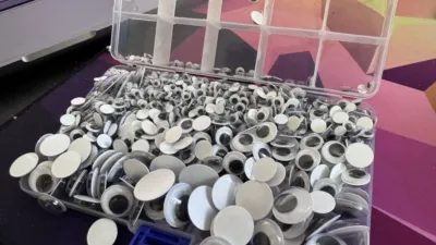 A no longer neatly sorted selection of googly eyes in various sizes, sitting on the lid of a compartmented storage box, with the compartments now vertically in the air.